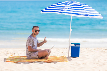 Happy young man in the beach under umbrella near the ocean working on his laptop and showing thumbs...