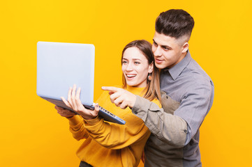 Portrait of happy cheerful couple looking and pointing at laptop over yellow background