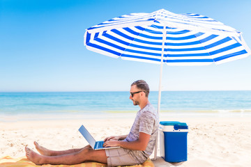 Young handsome man lusing laptop computer on beach bench with blue solar umbrella overhead, surrounded by turquoise sea and beautiful sky.
