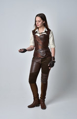 full length portrait of brunette  girl wearing brown leather steampunk outfit. standing pose on grey studio background.