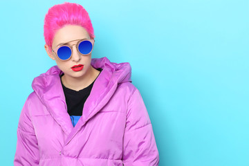Obraz premium Fashion portrait of young hipster woman in lilac down jacket. Short pink hair, sunglasses, red lips, turquoise background