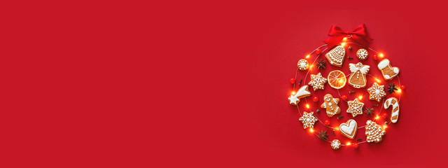Creative Christmas ball made of iced gingerbread cookies, anise stars, berries, orange chips, decorated red ribbon bow on red background. New Year banner. Top view.