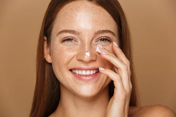Poster Beauty image of pretty shirtless woman smiling and applying face cream, isolated over beige background © Drobot Dean