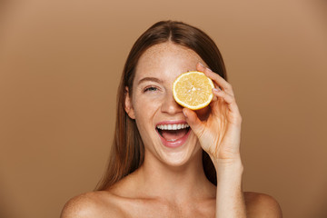 Beauty image of joyful shirtless woman with long hair holding piece of orange, isolated over beige background