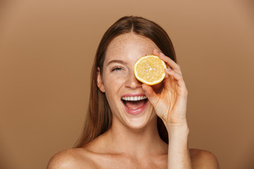 Beauty image of joyful shirtless woman with long hair holding piece of orange, isolated over beige...