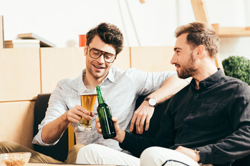 smiling friends clinking glass and bottle of beer in cafe