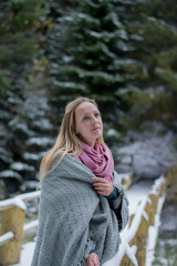  Girl near the winter lake wrapped in a blanket. Snowy forest and the bridge. Portrait of a blonde in nature