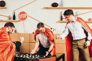 young friends in superhero costumes playing table soccer in cafe