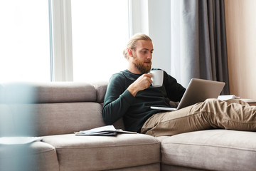 Concentrated young bearded man sitting in home using laptop computer.