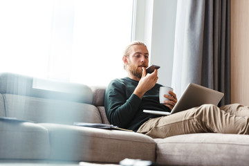 Young bearded man sitting in home using laptop computer and mobile phone.