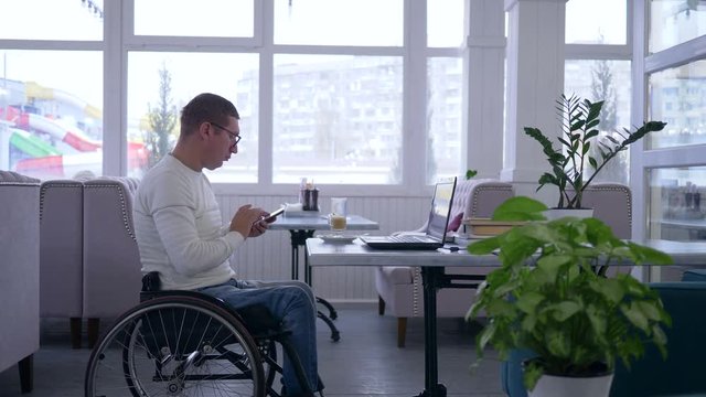 business freelance, senior man crippled in wheelchair wearing glasses uses a mobile phone and working on a laptop sitting at a table in a cafe