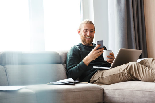 Young bearded man sitting in home using laptop computer and mobile phone.