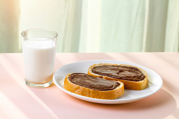 A pieces of loaf with spread chocolate paste on a white plate. Sweet, chocolate sandwiches with nut paste and a glass of milk for breakfast.