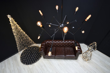 New year's Loft-style decor against a black wall, a Christmas tree from a garland. black chandelier with incandescent lamps and brown sofa. Beautiful New Year's decor with lighting in the studio