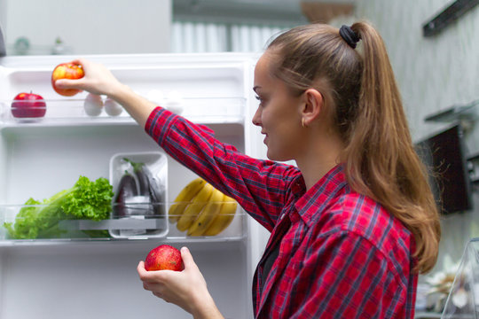Young, attractive girl pulls out a fresh apple for a snack from the fridge. Food storage, vegetables and fruits in the refrigerator. Proper, healthy and fresh food.