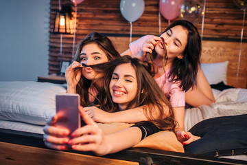 Three playful young women taking selfie on bed in room. Two models play with hair of third girl....