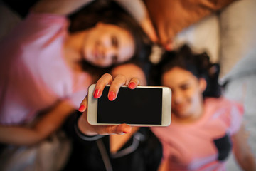 Picture of white phone in hand of one girl. She lies on bed with her friends. Models smile. They look happy.