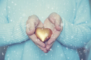 Woman hands are holding a beautiful glossy gold heart in a snow winter background. Love and St. Valentine cozy concept.