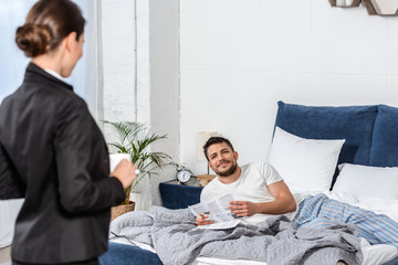 girlfriend holding cup of coffee in suit and boyfriend in pajamas reading newspaper in bedroom in...