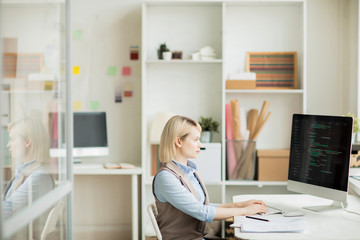 Serious concentrated beautiful lady with blond hair sitting at table and typing on computer keyboard while working with computer language