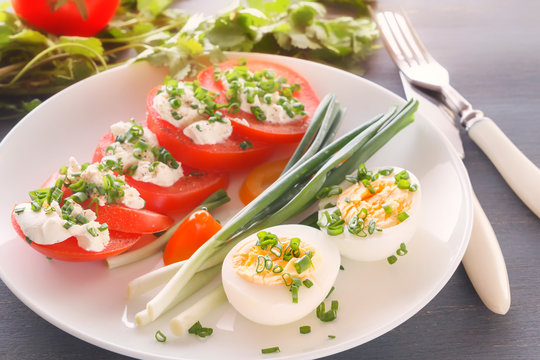 Half boiled eggs with tomatoes, green onions sprinkled with greens in a white plate on a gray wooden table. Close-up