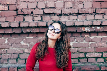 Fashionable young girl with long hair, red sweater in sunglasses. Model on brick wall background.
