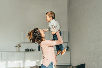 side view of smiling woman raising up adorable son in living room at home