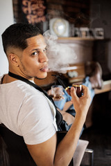 Young hispanic haidresser and hairstylist sitting in barber shop, smoking a pipe.