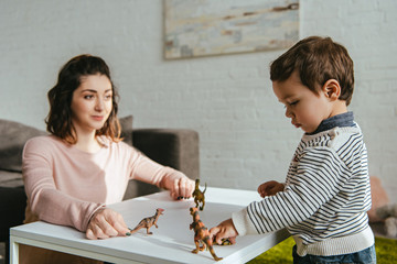 selective focus of mother and little son playing toy dinosaurs at table in living room at home