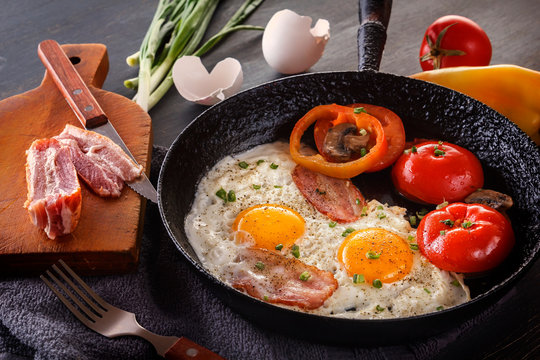 Fried eggs with bacon and tomatoes on an old cast iron pan and cutlery on a gray table. Close-up