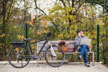 Obraz premium Senior man with electrobike sitting on a bench outdoors in town, using laptop and smartphone.
