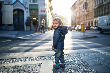 A small toddler boy standing by a road outdoors in city at sunset, looking back. - Powered by Adobe