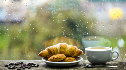 Coffee time on rainy day. Fresh brewed coffee in white cup or mug on windowsill. Wet glass window and cup of hot caffeine beverage. Coffee drink with croissant dessert. Enjoying coffee on rainy day