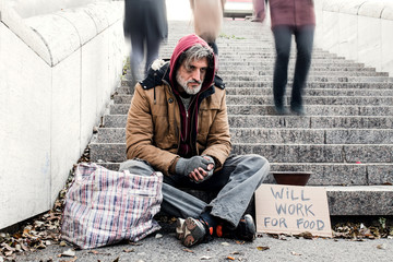 Homeless beggar man sitting outdoors in city asking for money donation.