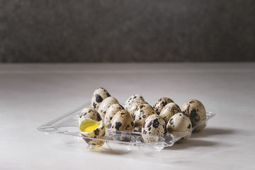 Raw uncooked quail eggs whole and broken in plastic boxing on white marble kitchen table.