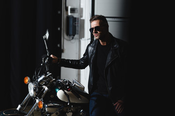 stylish man in black sunglasses posing by motorbike in garage