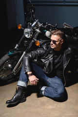 Fototapeta na wymiar handsome biker in sunglasses sitting by motorcycle in garage
