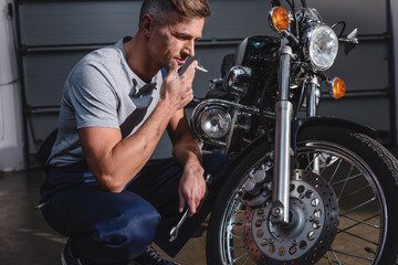Fototapeta na wymiar handsome adult mechanic smoking and holding wrench while fixing motorcycle in garage