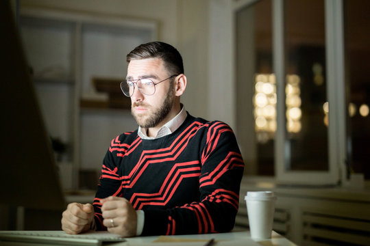 Serious exhausted young male manager in glasses frowning forehead trying to not sleep while working at night in dark office