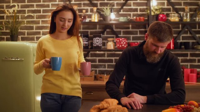 Displeased man having unpleasant taste of coffee made by callow young beautiful wife while couple having breakfast in domestic kitchen. Man drinking bitter coffee, expressing disgust and displeasure.