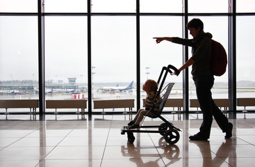 Silhouette of mother with her toddler son in stroller against the window at the airport. Mom points...