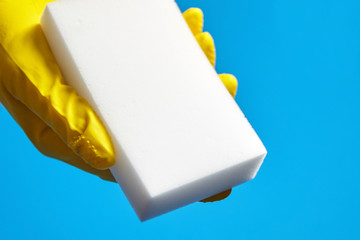 Human hand in yellow glove holds a white melamine sponge on blue background. Universal tool for cleaning various surfaces from dirt and stains