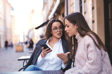 two beautiful stylish women sitting at the table in street cafe, looking at the phone and talking. women spending time together at bright sunny spring day and discussing something - 237020987