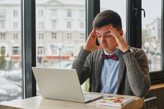 Young tired, ill, overworked man in formal wear sitting in front of computer and touching his forehead in the cafe