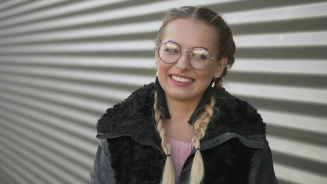 Slow motion portrait of beautiful young blonde woman in glasses, braids hairstyle, looking down, then into camera and smiling. Posing against urban metal strips background.