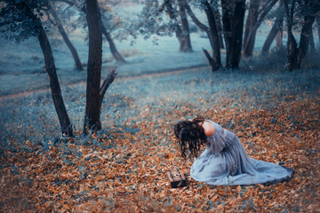 dark-haired girl in a long, light gray, old-fashioned dress, sits on cold ground covered with...