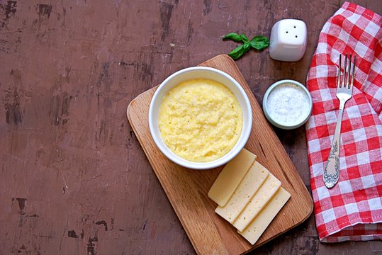 Polenta or hominy, a dish of corn grits or corn flour, in a white bowl on a brown background. Served with sour cream and mint sauce and cheese. Top view.