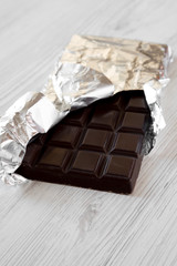 Dark chocolate in a wrapper over white wooden background, side view.
