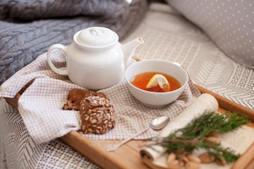 Fototapeta na wymiar White teapot, mug of lemon tea, rye bread with seeds are serving on wooden tray on sofa. Morning breakfast, hot beverage in cozy home interior. Autumn or Christmas mood.