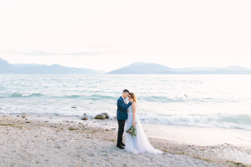 Tender portrait of young man hugging his pretty woman while standing on the pier near the lake on the wedding ceremony. Garda Lake, Italy